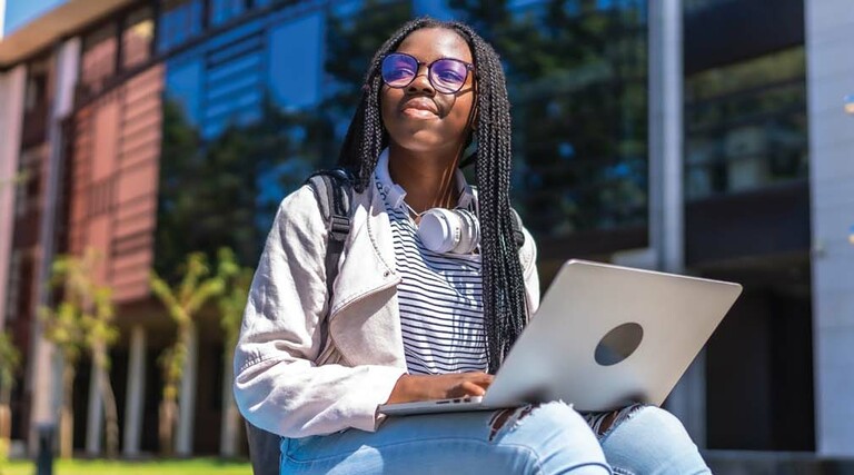 A college student with a laptop on her lap gazes skyward outside a campus building